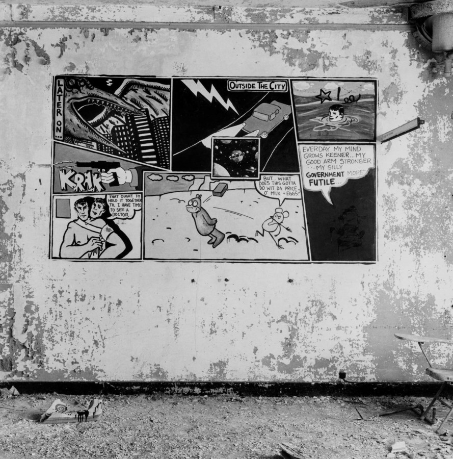 Black-and-white photograph of a derelict interior with comic panels painted on the wall