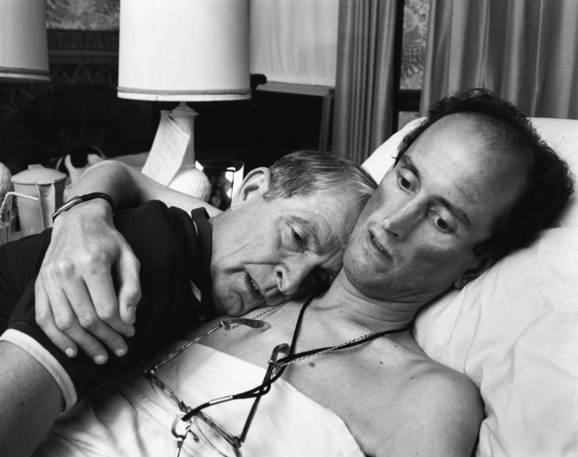 Black-and-white photograph of a young man in bed embracing an older man on his shoulder with one arm