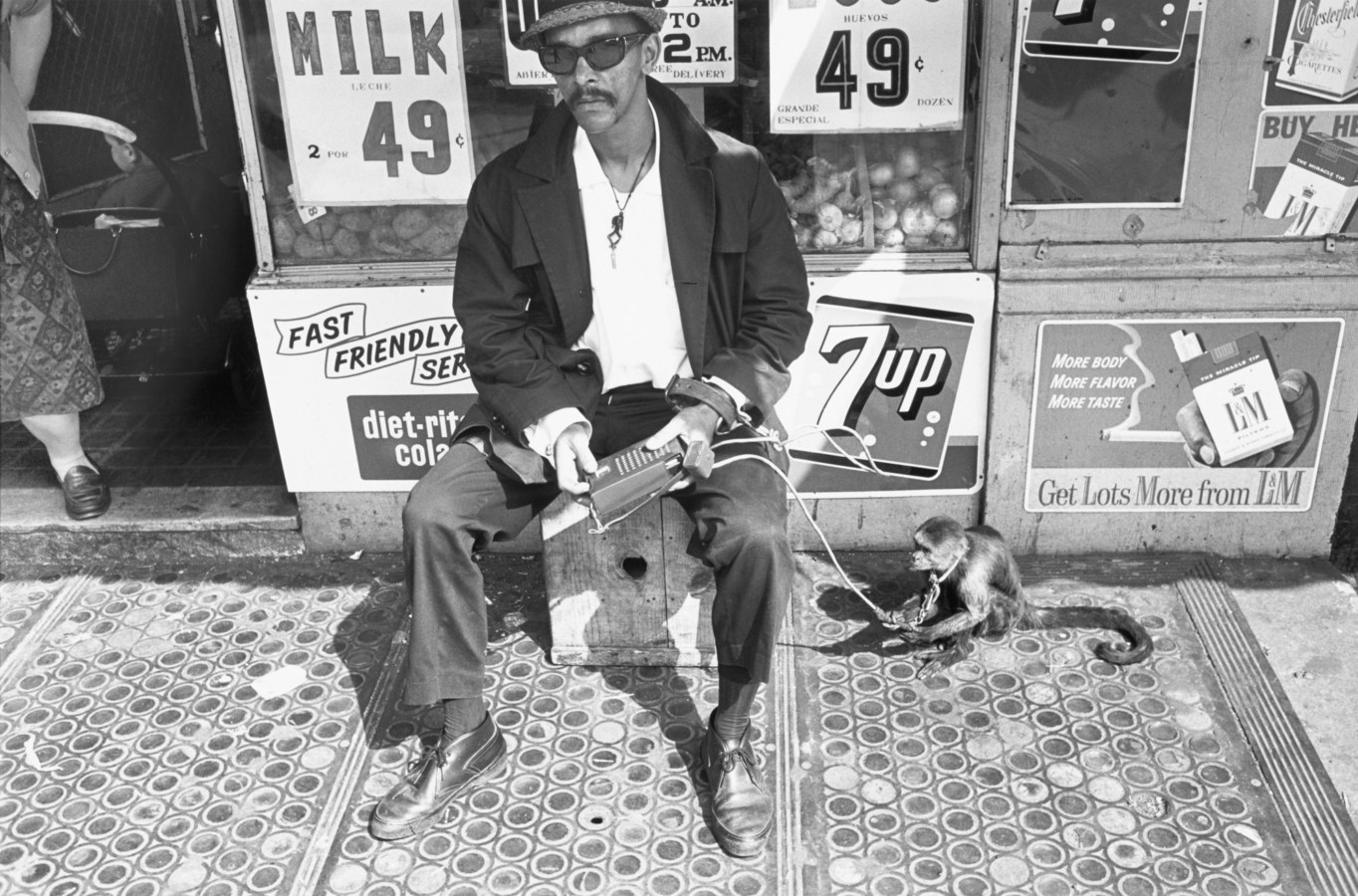 Black-and-white photograph of a man and a monkey sitting in front of a store with advertisements in the background