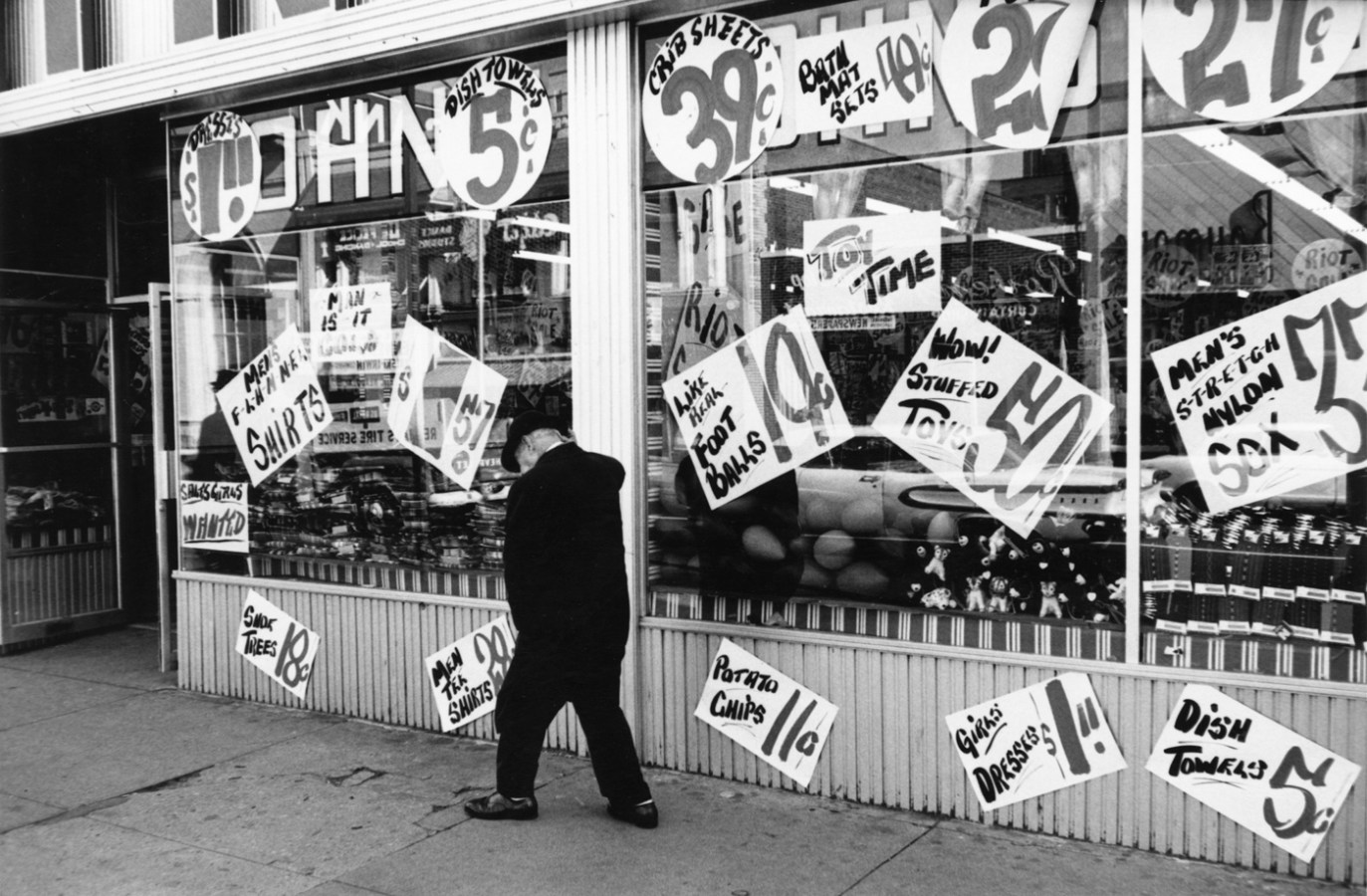 Black-and-white photograph of a man walking in in front of a storefront covered in hand painted signs advertising the prices of products