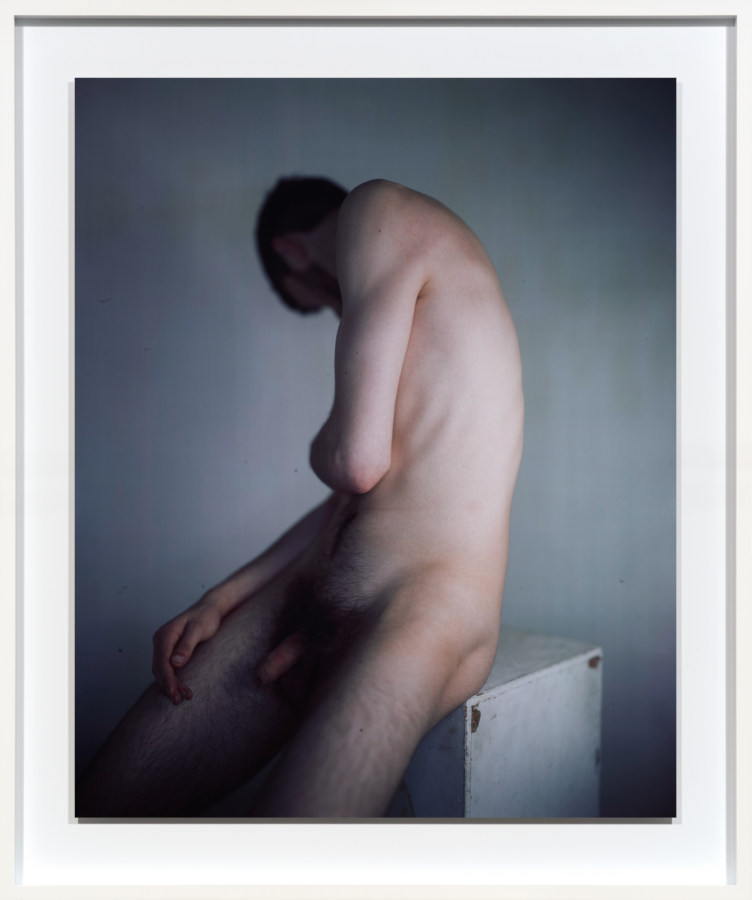 Color photographic portrait of a nude man seated on a plinth with his face turned to his shoulder away from the camera