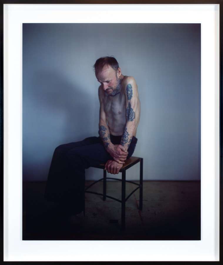 Color photographic portrait of a seated shirtless man with tattooed arms looking downwards
