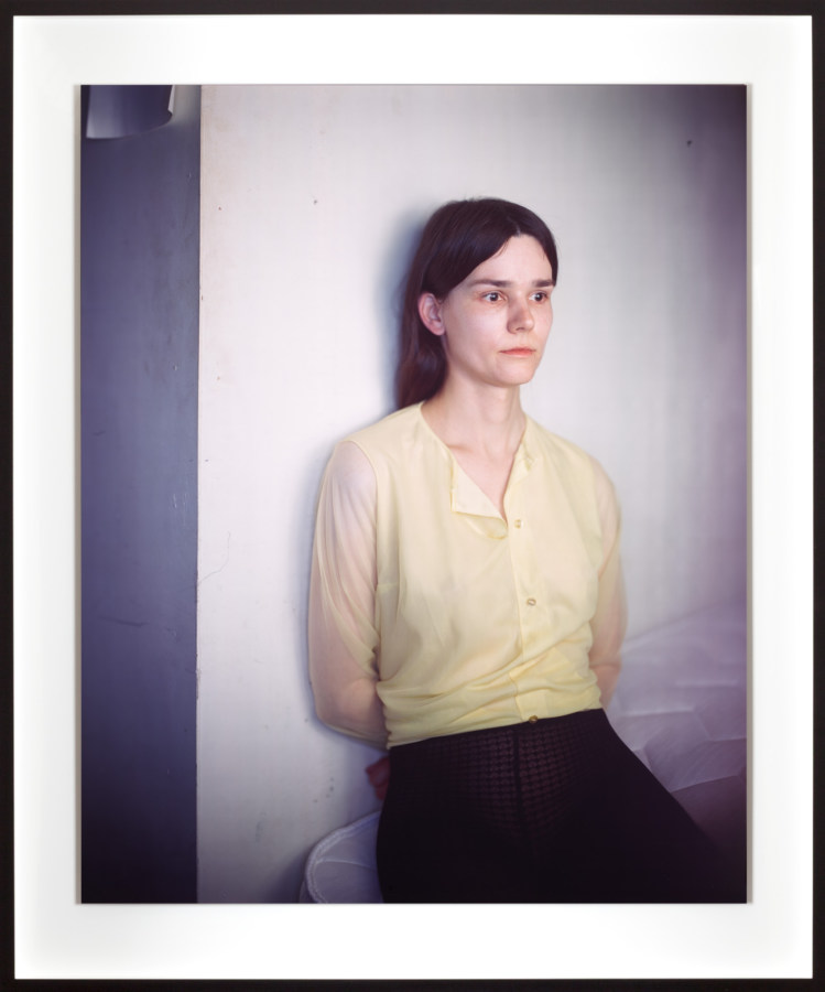 Color photographic portrait of a seated young woman in a yellow shirt leaning on a wall with her arms behind her back