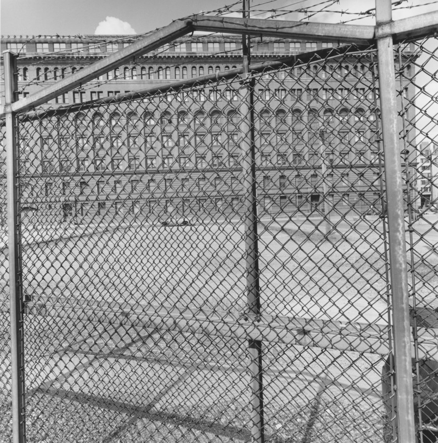 A black and white photograph of a chain link fence with a large structure in the background