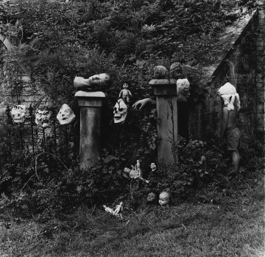 Black-and-white photograph of an assortment of rubber masks and doll heads positioned on two pedestals in an overgrown garden