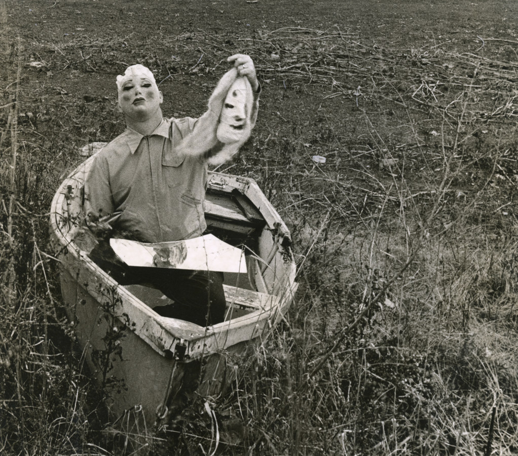 Black-and-white photograph of a person wearing a rubber mask seated in a rowboat on the grass holding up another mask