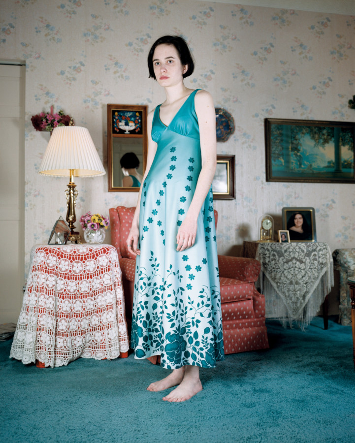 Color photograph of a girl in a long teal flowered dress standing in front of a red armchair in a room with flowered wallpaper