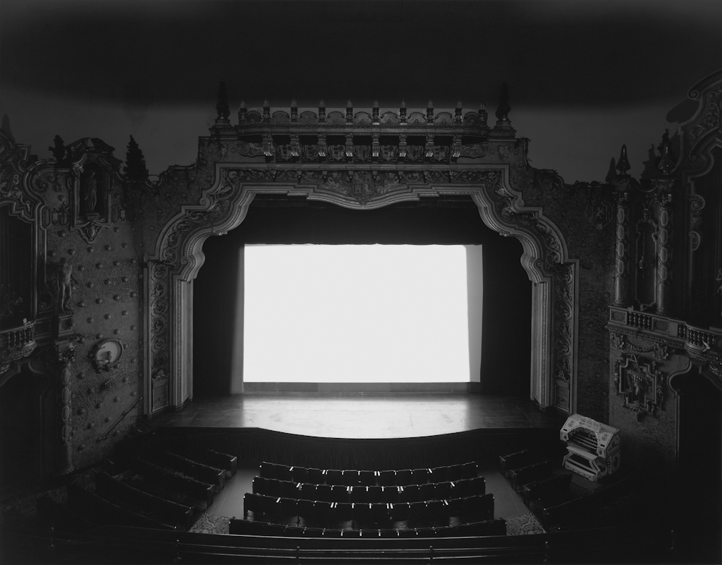 Black-and-white photograph of an empty theater with a glowing white screen onstage