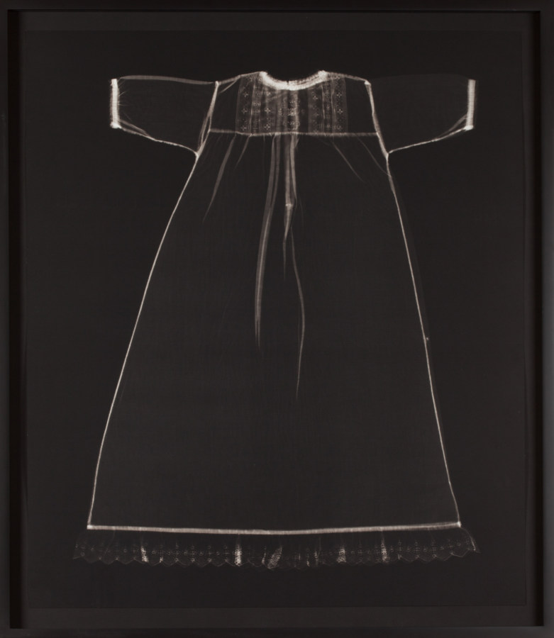 Black-and-white photograph of the faint white outline of a child's short-sleeved dress on a black background