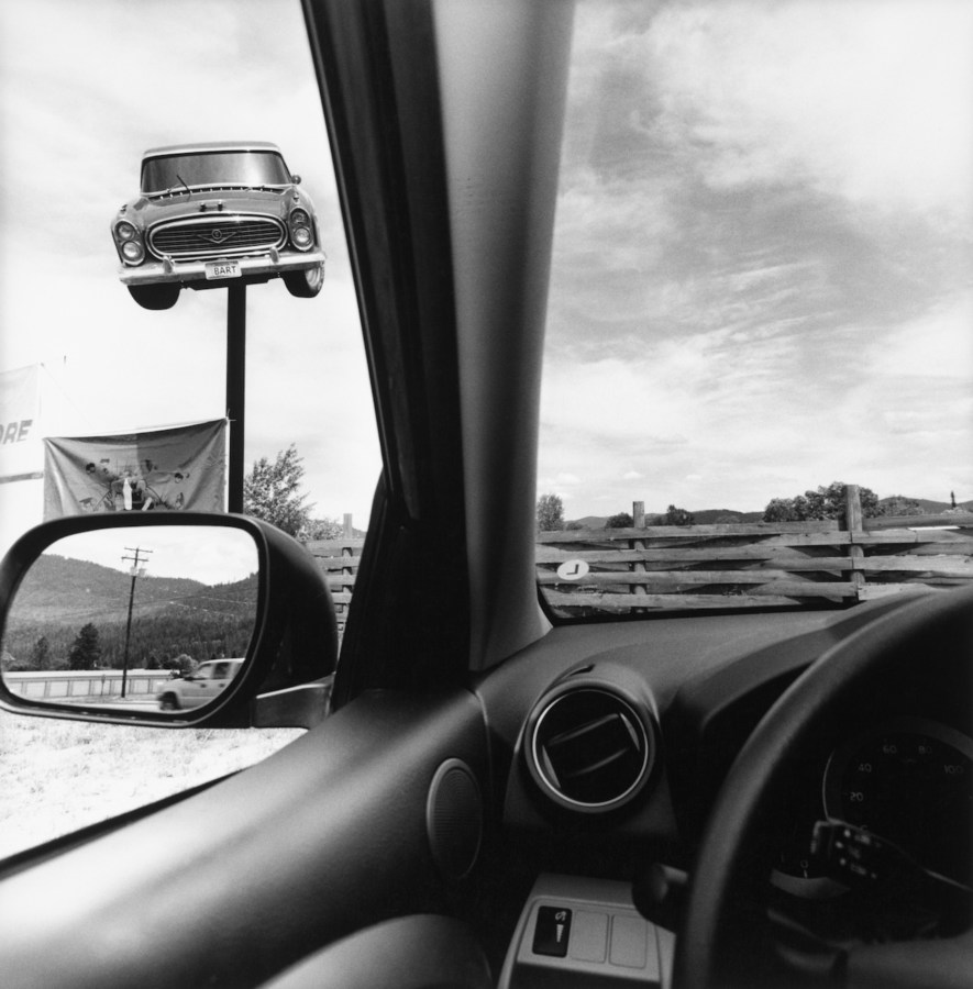 Black-and-white photograph from the drivers seat of a car showing the interior of the car and an advertisement of a 1950s car mounted on top of a pole