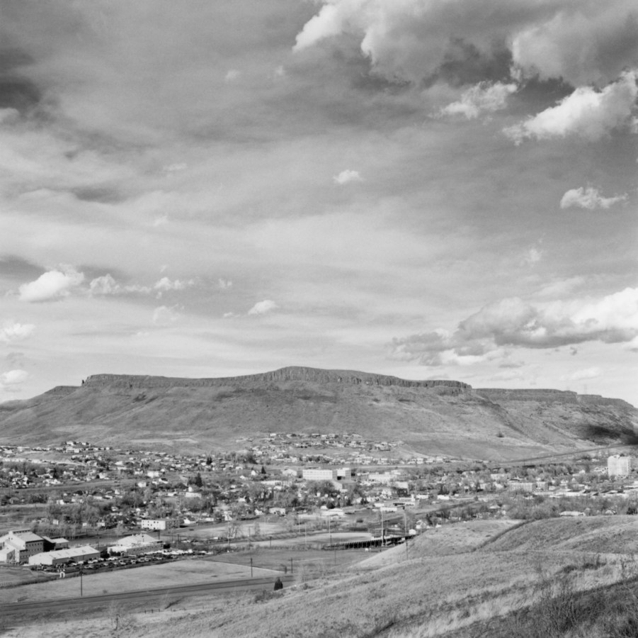 A black and white photograph overlooking a small town with mountains and clouds in the distance.