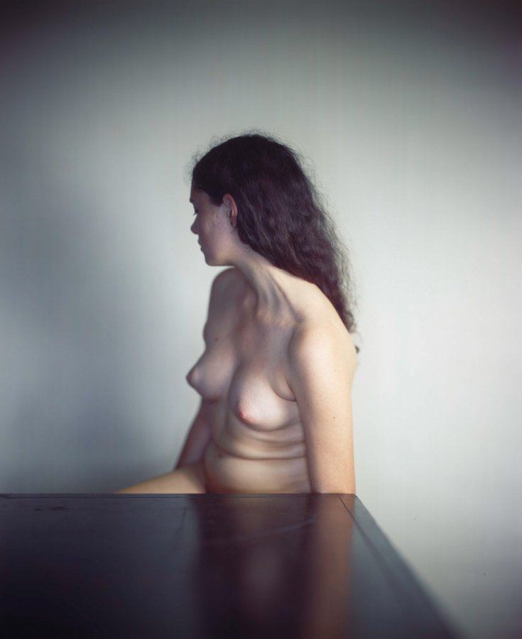Color photograph of a nude white woman seated behind a table, turned away from the camera.