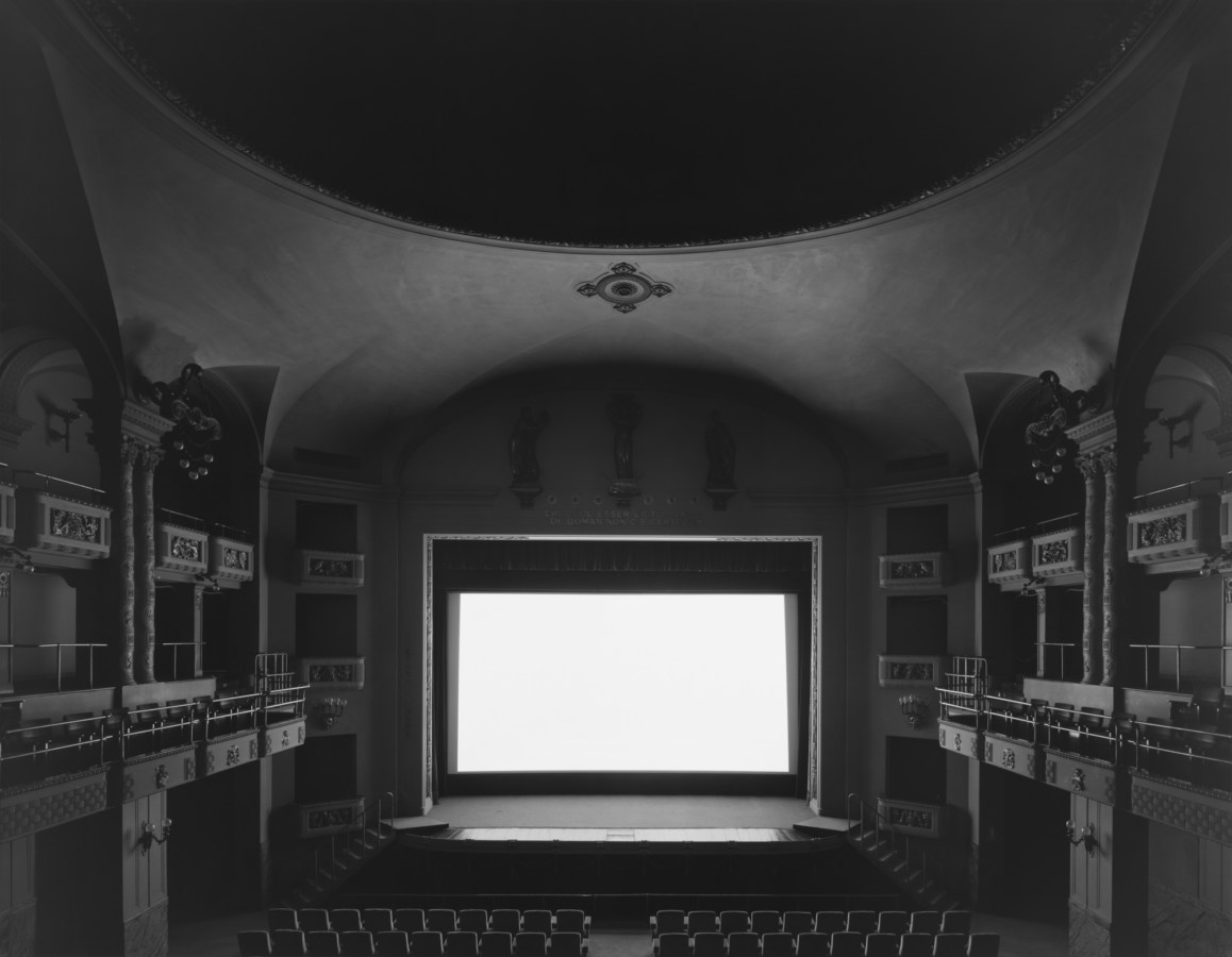 Black-and-white photograph of an empty theater with a glowing white screen onstage