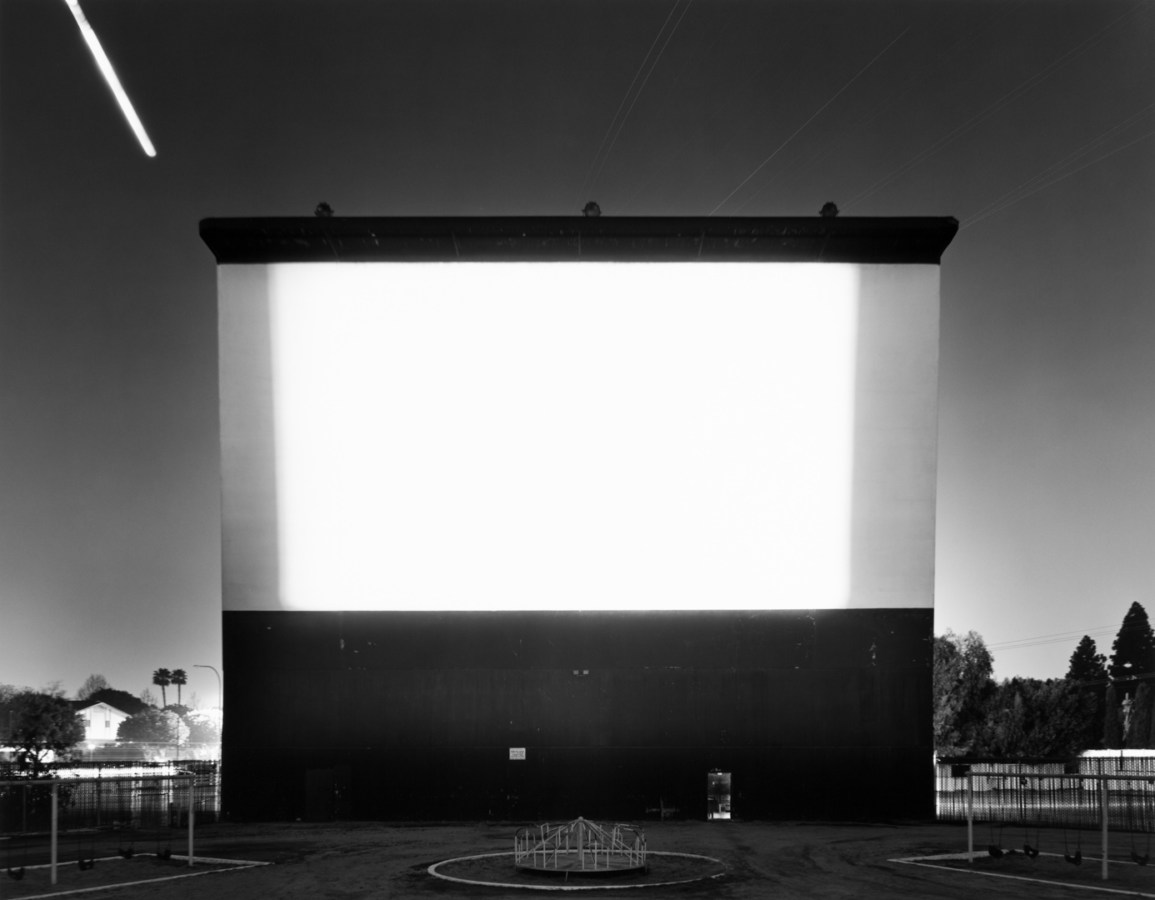 Black-and-white photograph of a blank white outdoor movie screen against an American suburb