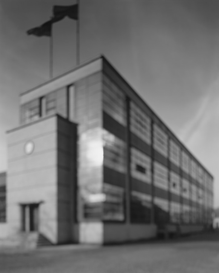 Black-and-white photograph of out of focus factory building with rows of large glass windows and two flags on the roof