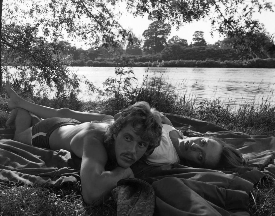 Black-and-white photograph of a man and a woman lying on a blanket next to a riverbank