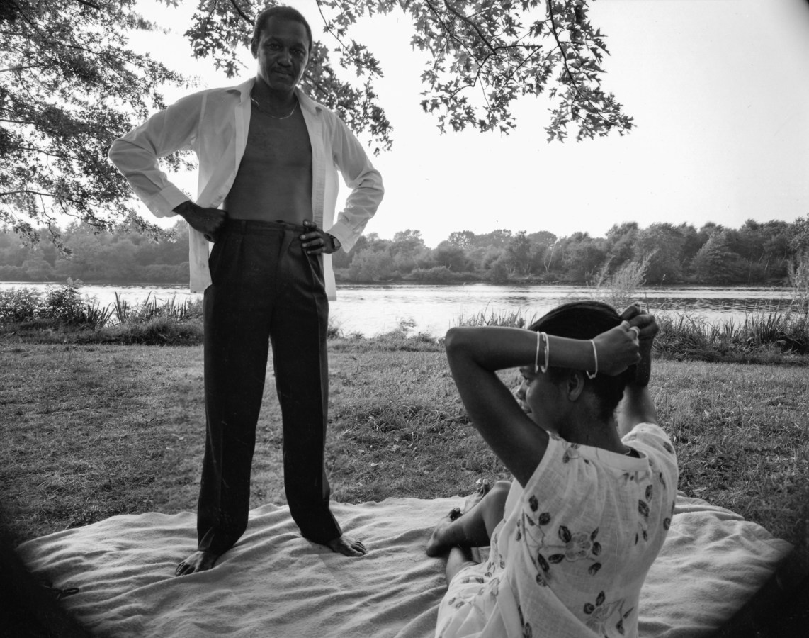 Black-and-white photograph of a man standing with his hands on his hips on a picnic blanket over a woman putting up her hair