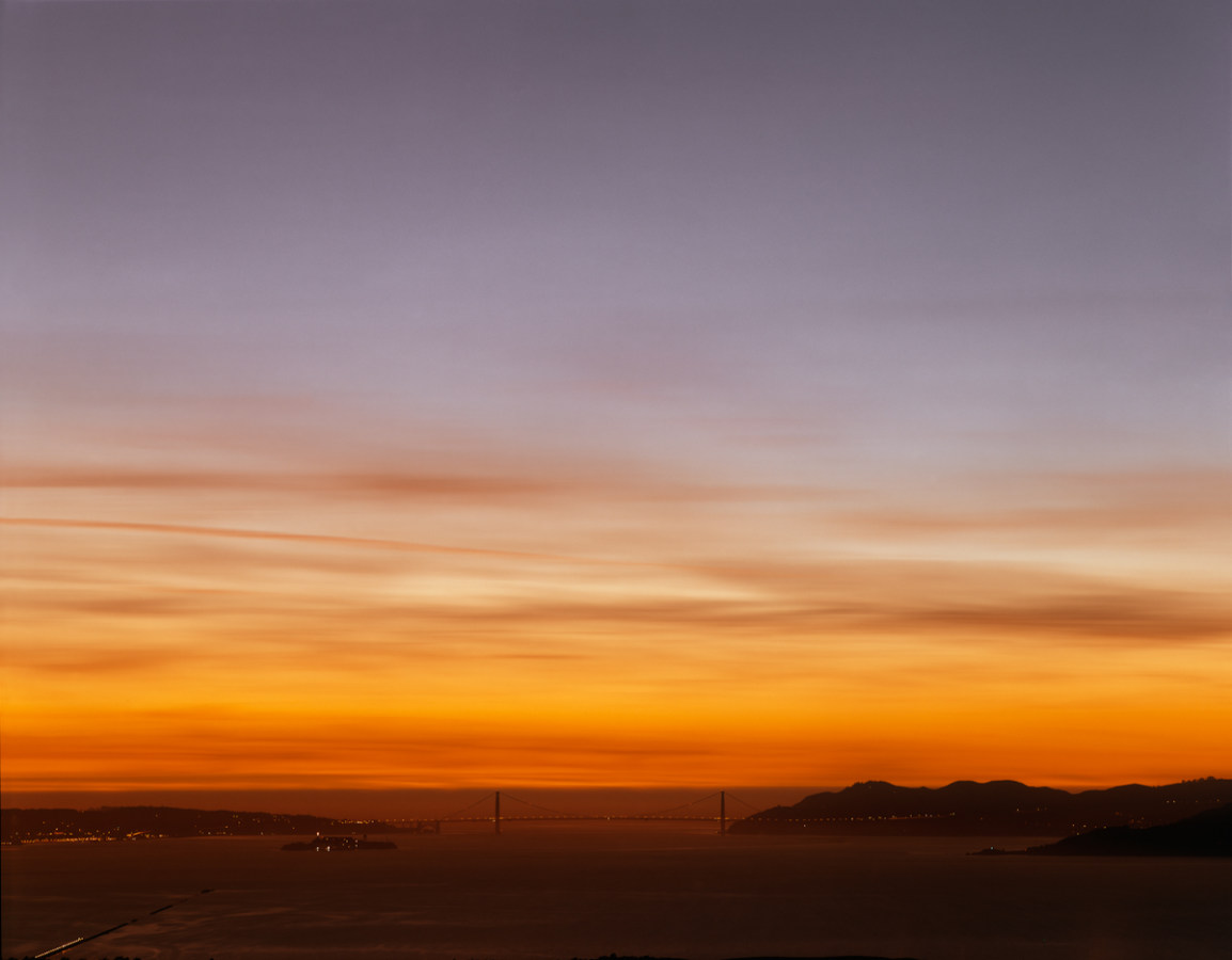 Color photograph of the Golden Gate Bridge on the horizon under a bright red-orange and violet sunset