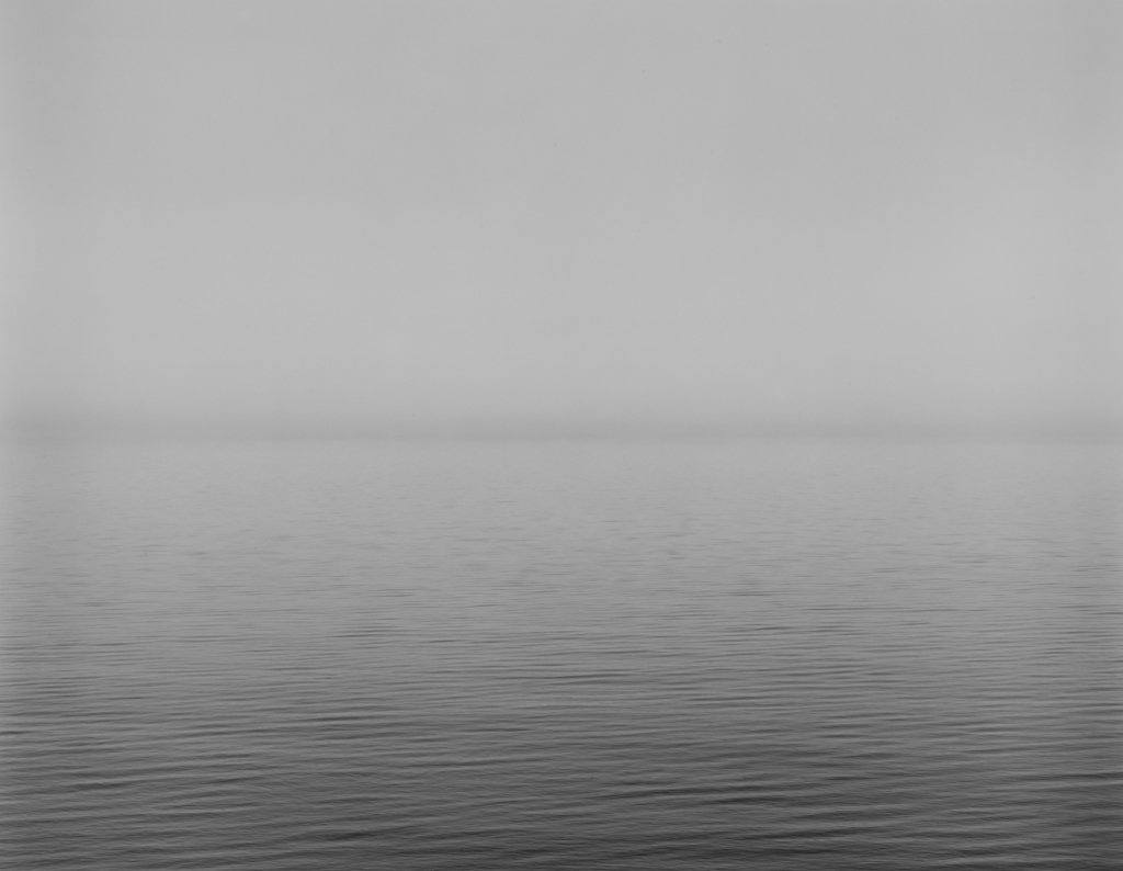 Black-and-white photograph of a calm waterscape across the lower half with a foggy sky above