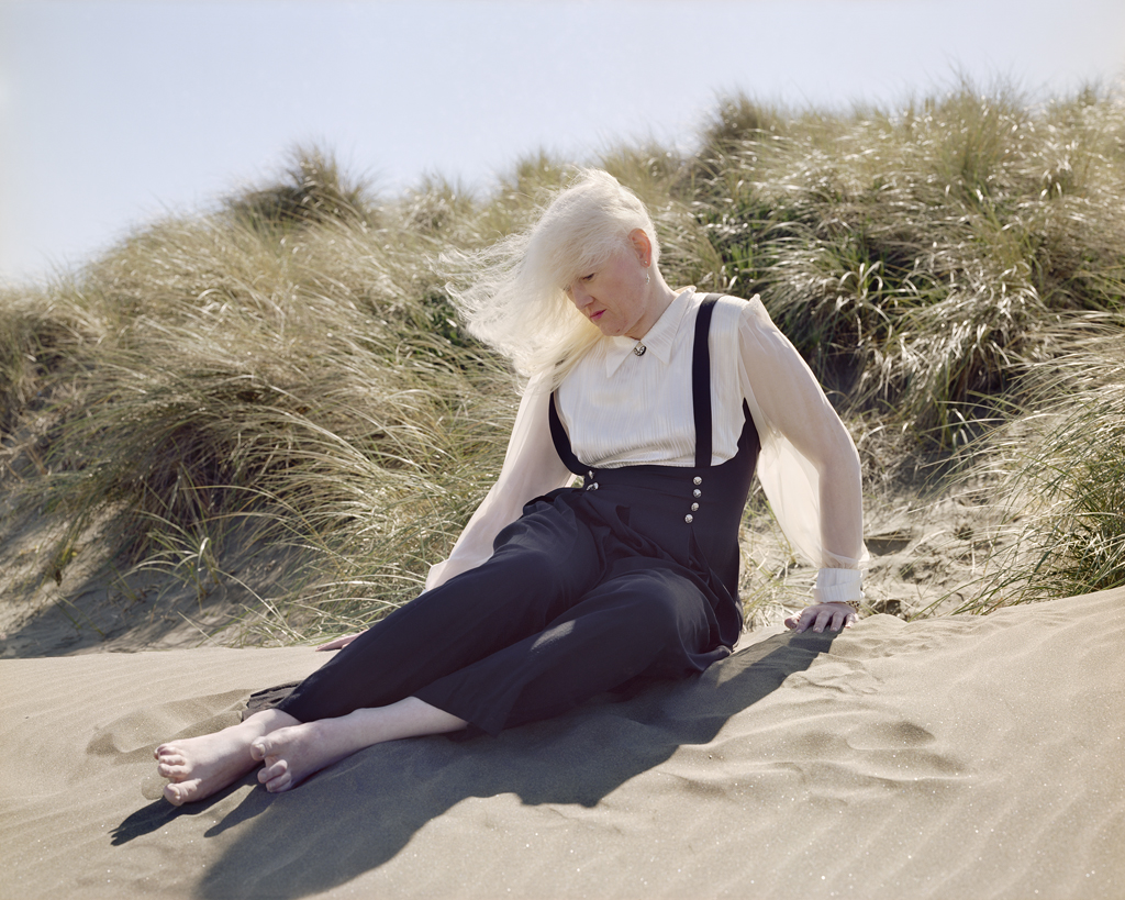 Color photograph of a woman with long white hair blowing in the wind seated on a sand dune covered with beach grasses