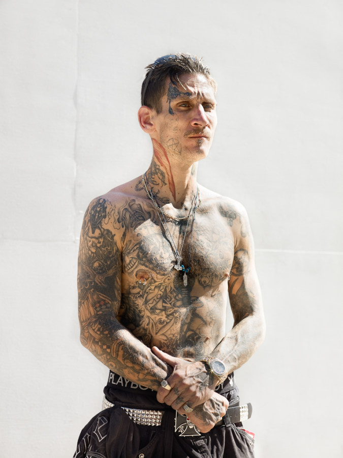 Color photograph of a shirtless tattooed man standing in front of a blank white wall