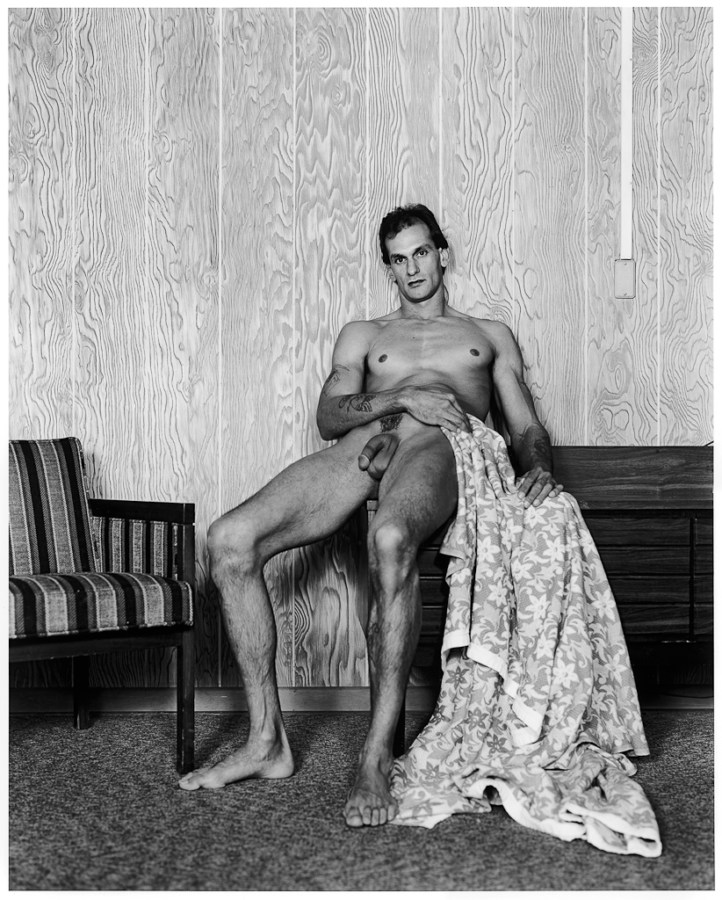 Black-and-white photograph of a nude man leaning against a wood paneled wall with a flowered blanket draped by his side