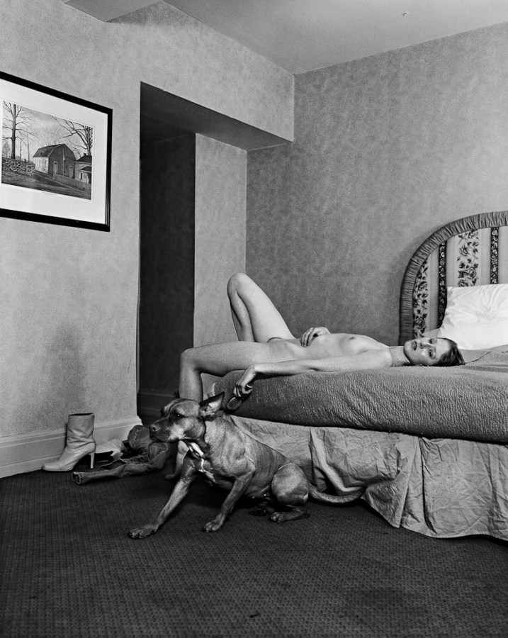 Black-and-white photograph of a nude woman lying on a bed with a dog seated on the floor beside her