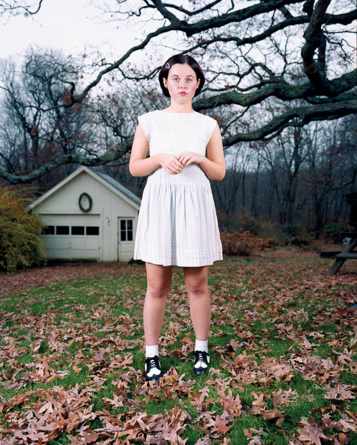Color photograph of a girl in a white dress standing on a lawn covered with fallen leaves under bare trees and a garage in the background