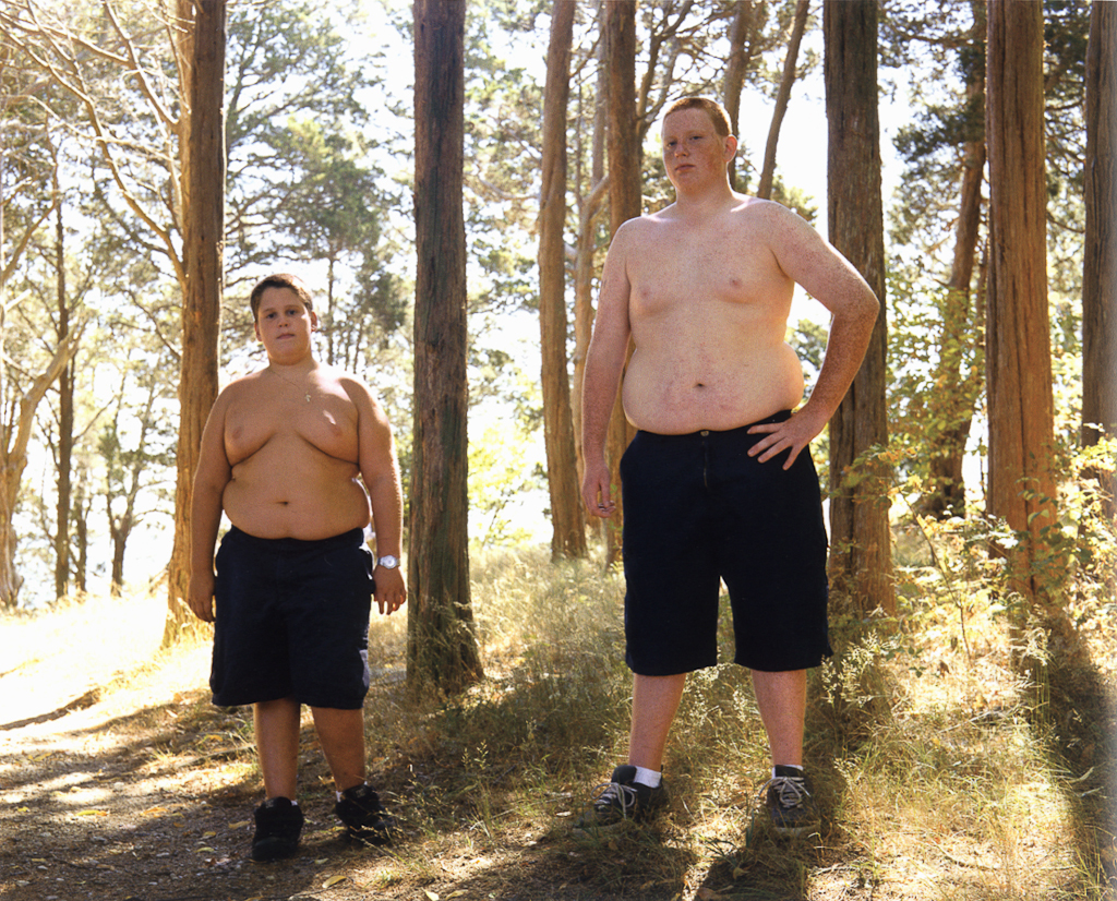 Color photograph of two boys wearing only shorts and sneakers standing in a sunny grove of trees