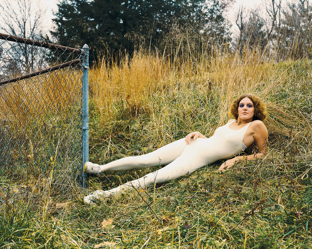 Color photograph of a woman in a white bodysuit reclining on a grassy hillside next to the corner of a chainlink fence