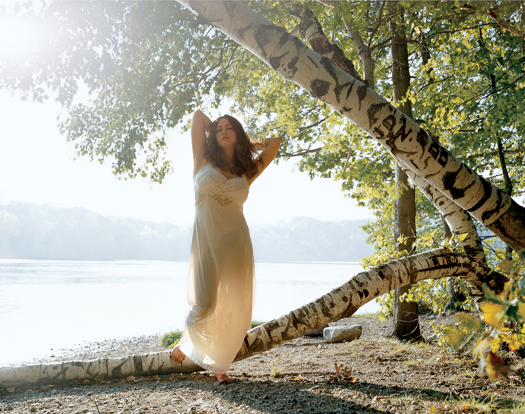 Color photograph of a woman in a long white dress standing on a fallen tree trunk on the shore of a still lake