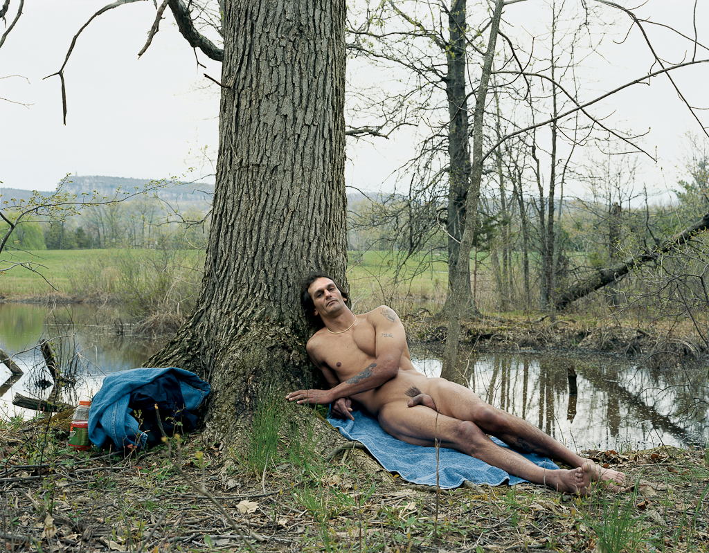 Color photograph of a nude man reclining against a tree next to a stagnant pond