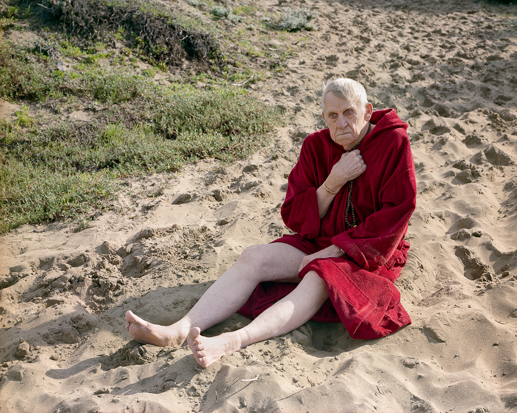 Color photograph of an elderly man seated on a sandy beach clutching a string of wooden beads and wrapped in a red towel