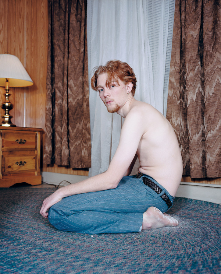 Color photograph of a shirtless young man kneeling on a rug in a wood paneled room
