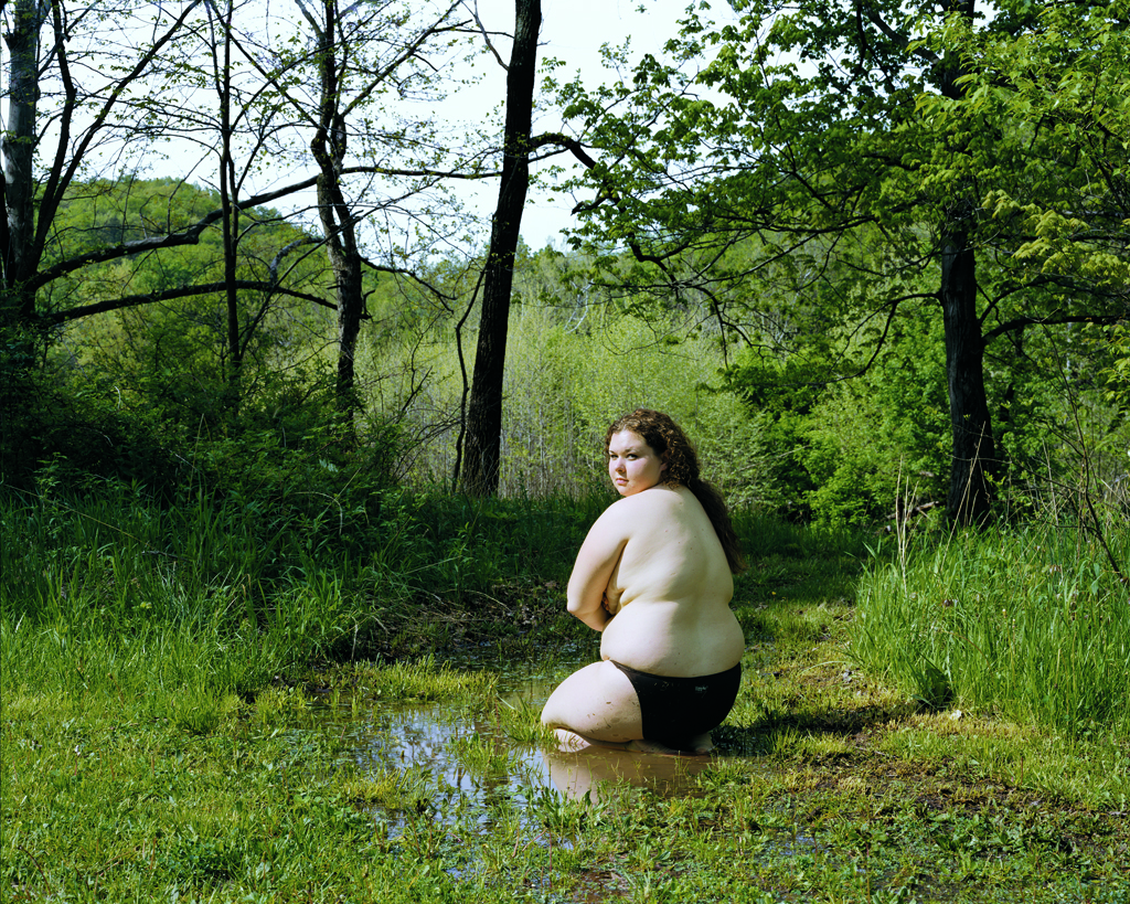Color photograph of a shirtless girl kneeling in a stagnant pool in a grassy clearing