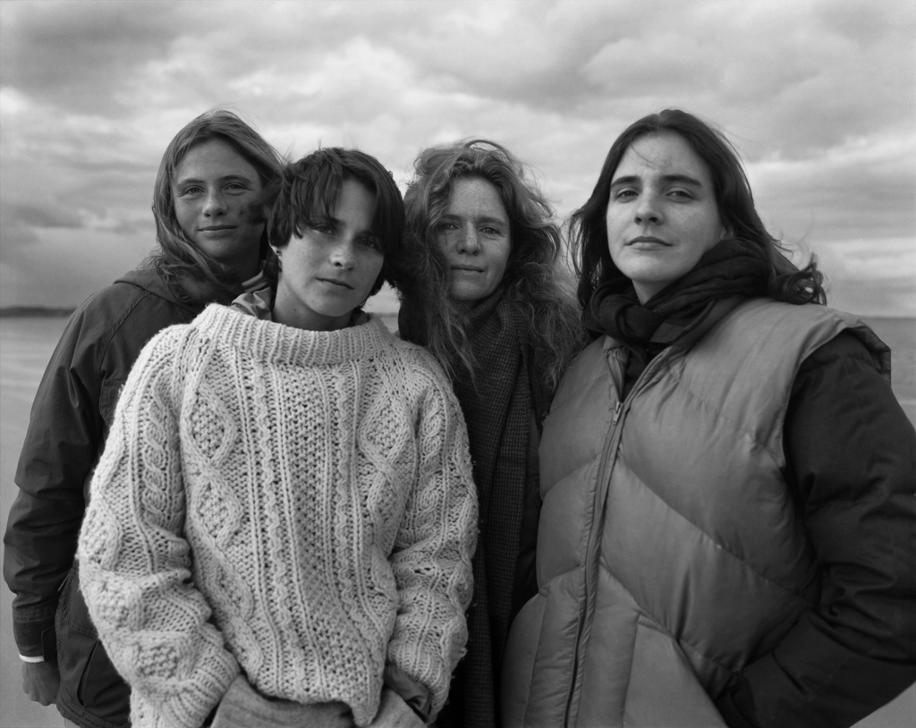 Black-and-white photographic portrait of four young women dressed in sweaters standing under a cloudy sky