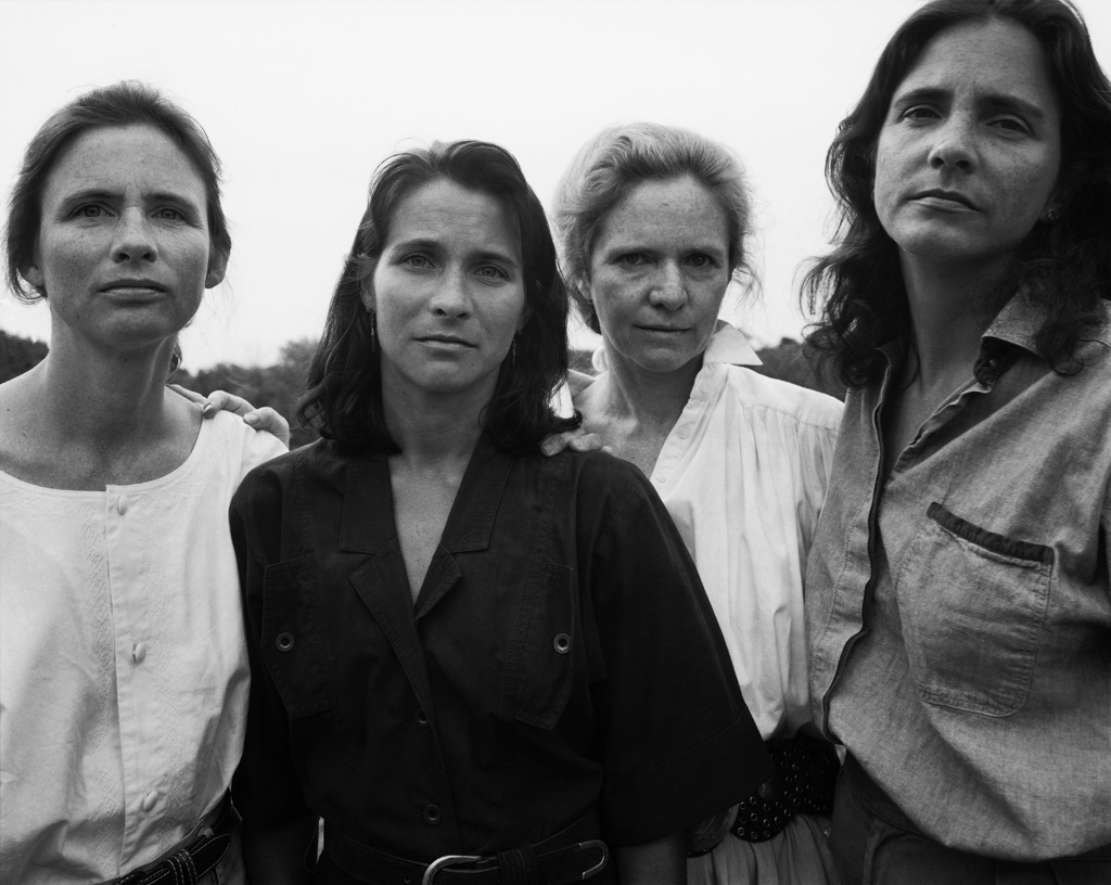 Black-and-white photographic portrait of four women standing looking slightly down at the viewer