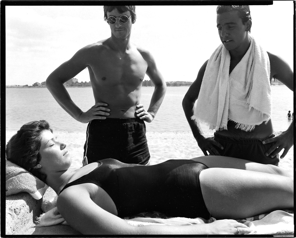 Black-and-white photograph of two young men in swimming shorts standing over a young woman sunbathing in a bathing suit lying down with her eyes closed