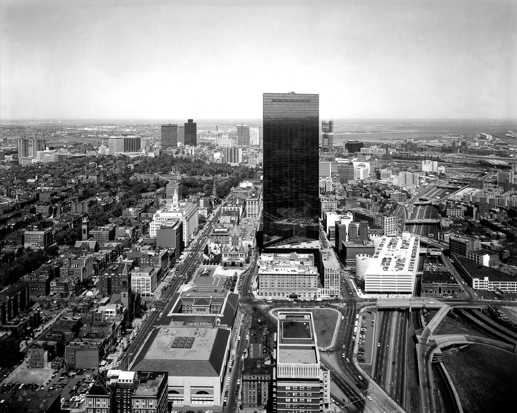 Black-and-white photograph of a rectangular glass high-rise building rising above surrounding city buildings