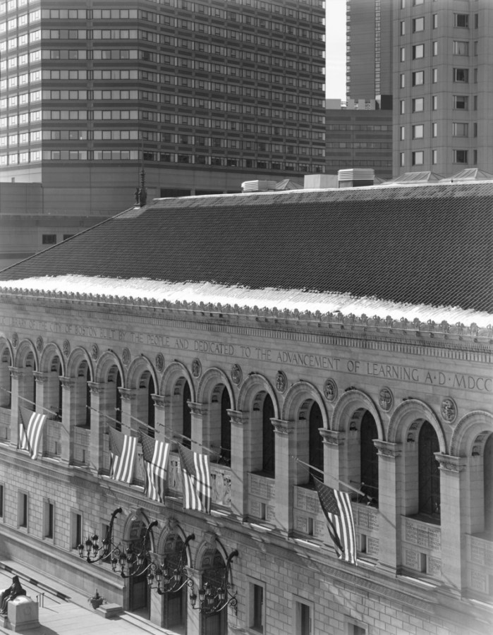 Black-and-white photograph of a low stone building flying American flags on its façade in front of modern high-rise buildings