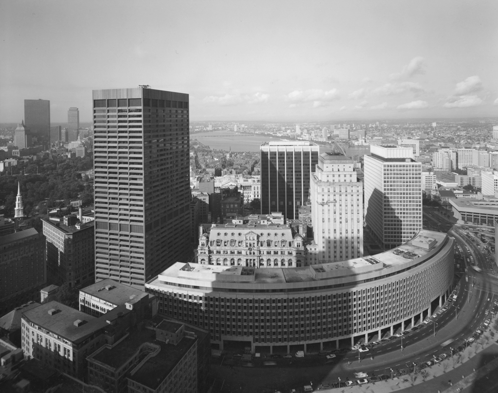 Black-and-white photograph of a curved building amid taller rectangular high-rise buildings