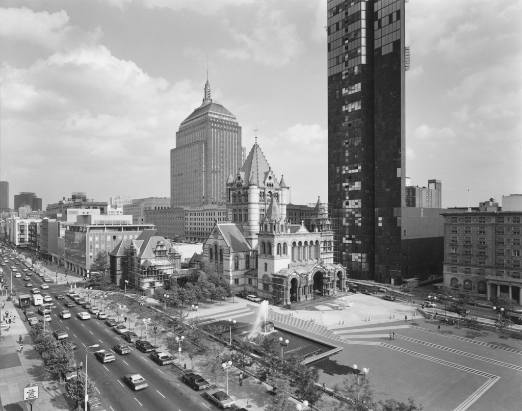 Black-and-white photograph of a large church set behind an open plaza amid modern city high-rise buildings