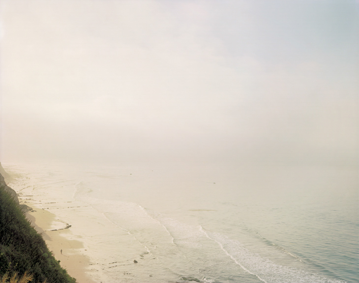 Color photograph of a lone figure walking along a beach shrouded in mist