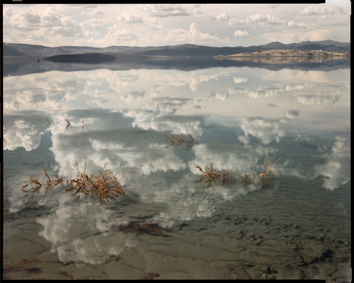 Color photograph of a still lake reflecting a cloudy sky and low mountains on the horizon