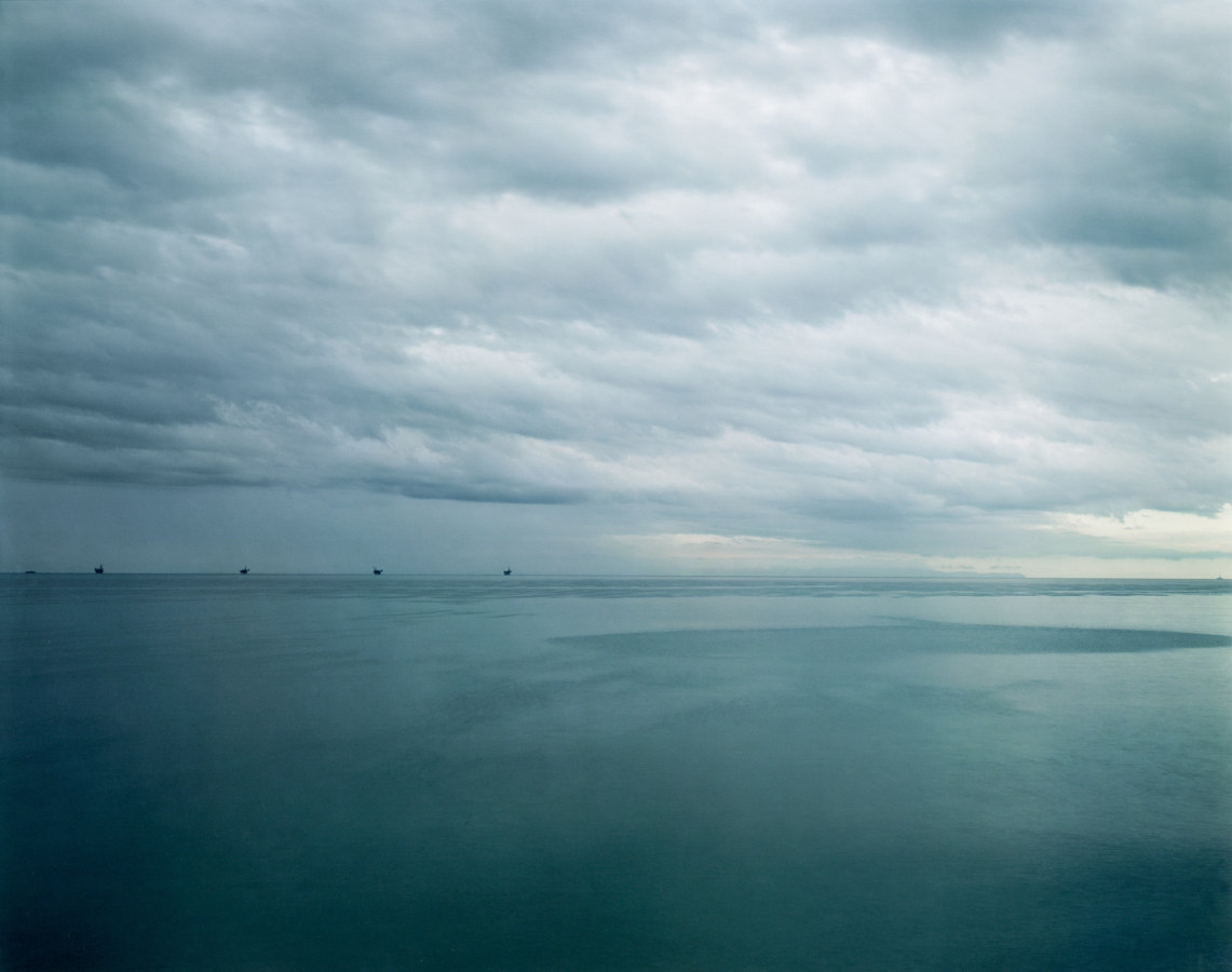 Color photograph of five black structures on the horizon rising above the waters of a calm sea