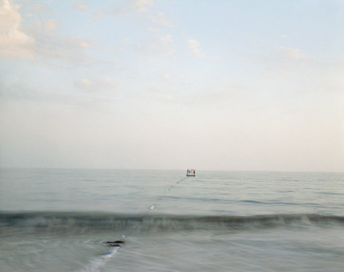 Color photograph of four people drifting towards the horizon on a raft on a calm sea