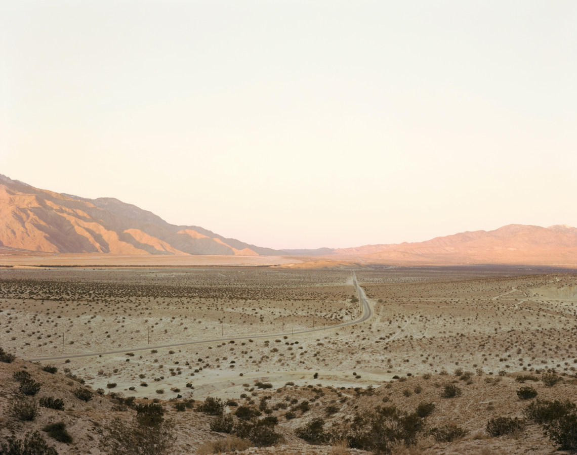 Color photograph of a road through a desert valley with mountains on the horizon