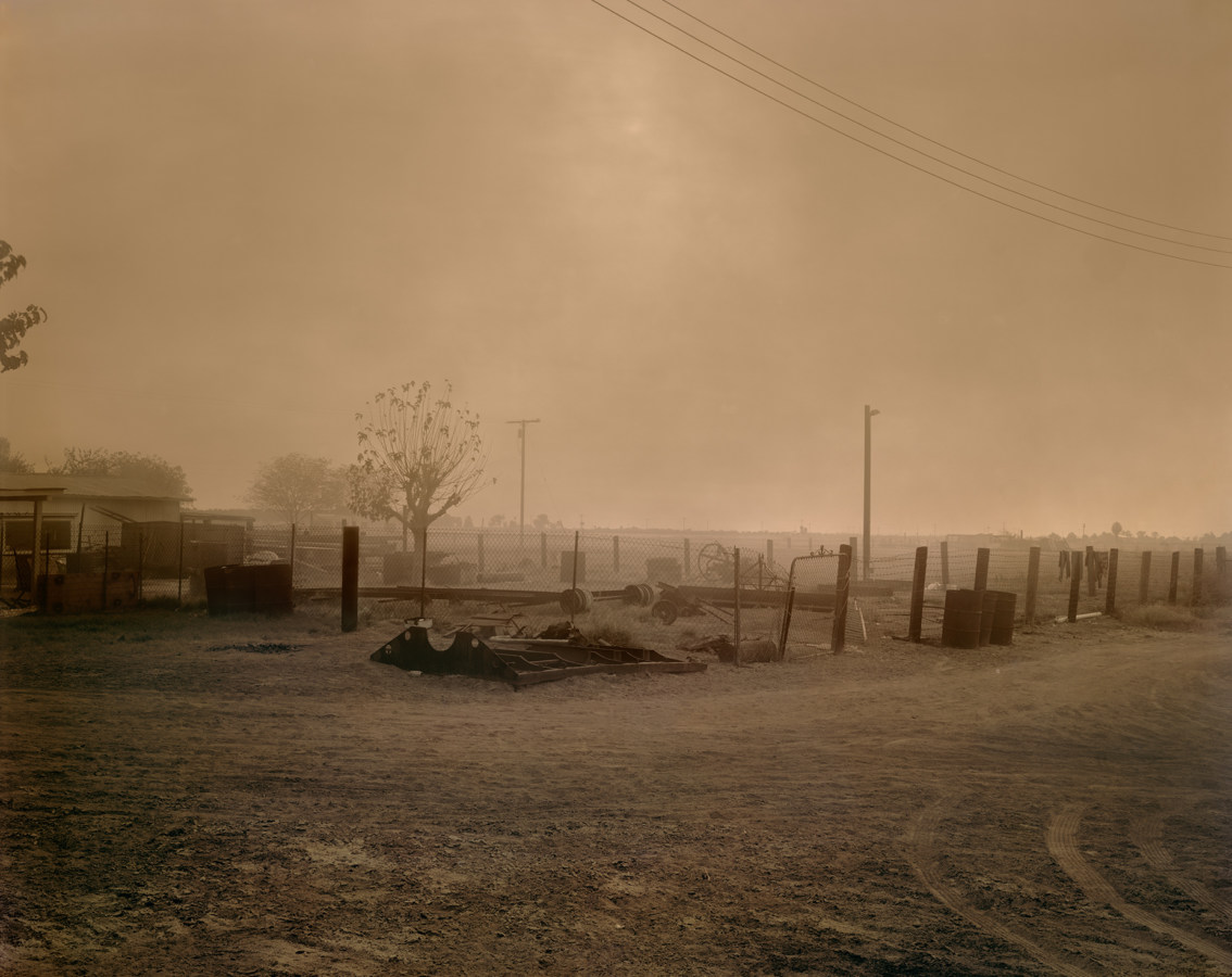 Color photograph of a smoke-filled sky above a fenced-in house and fields