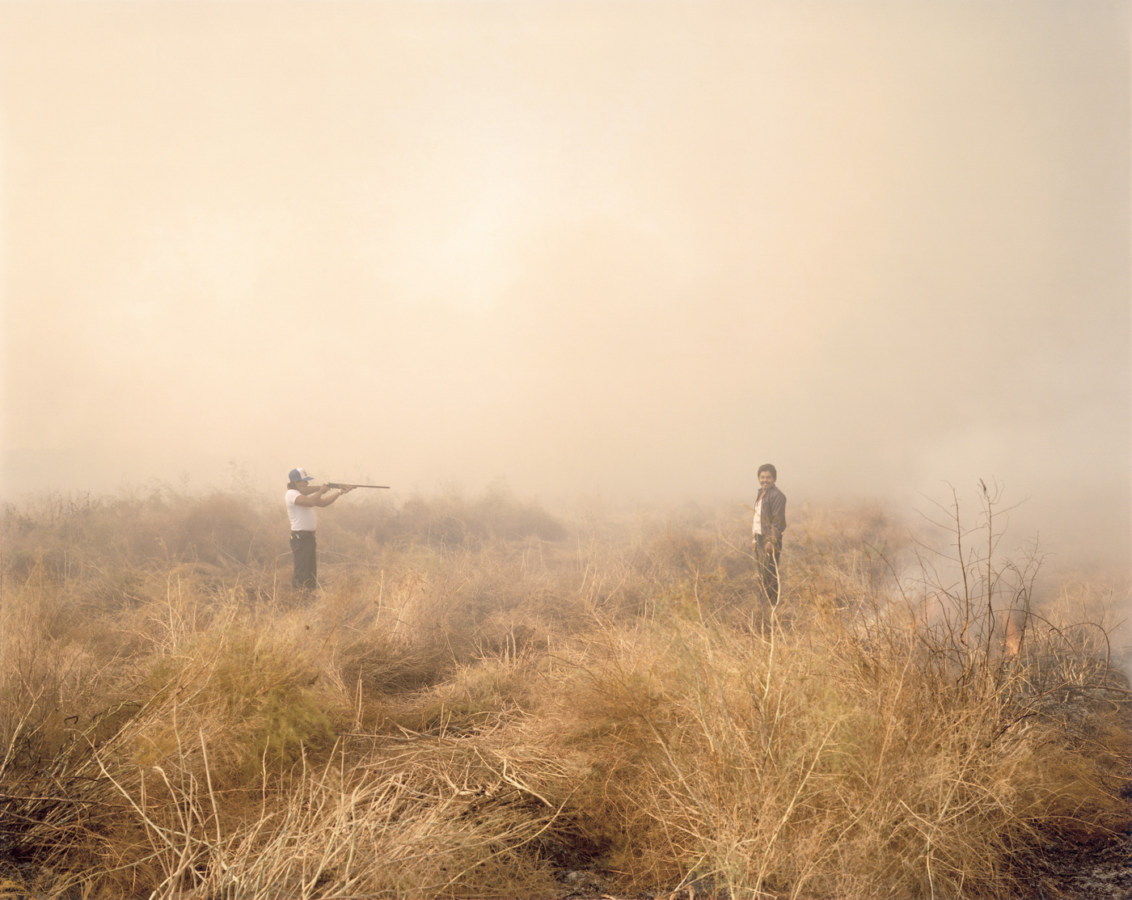 Color photograph of a man pointing a rifle at another man, both standing in a smokey scrubland