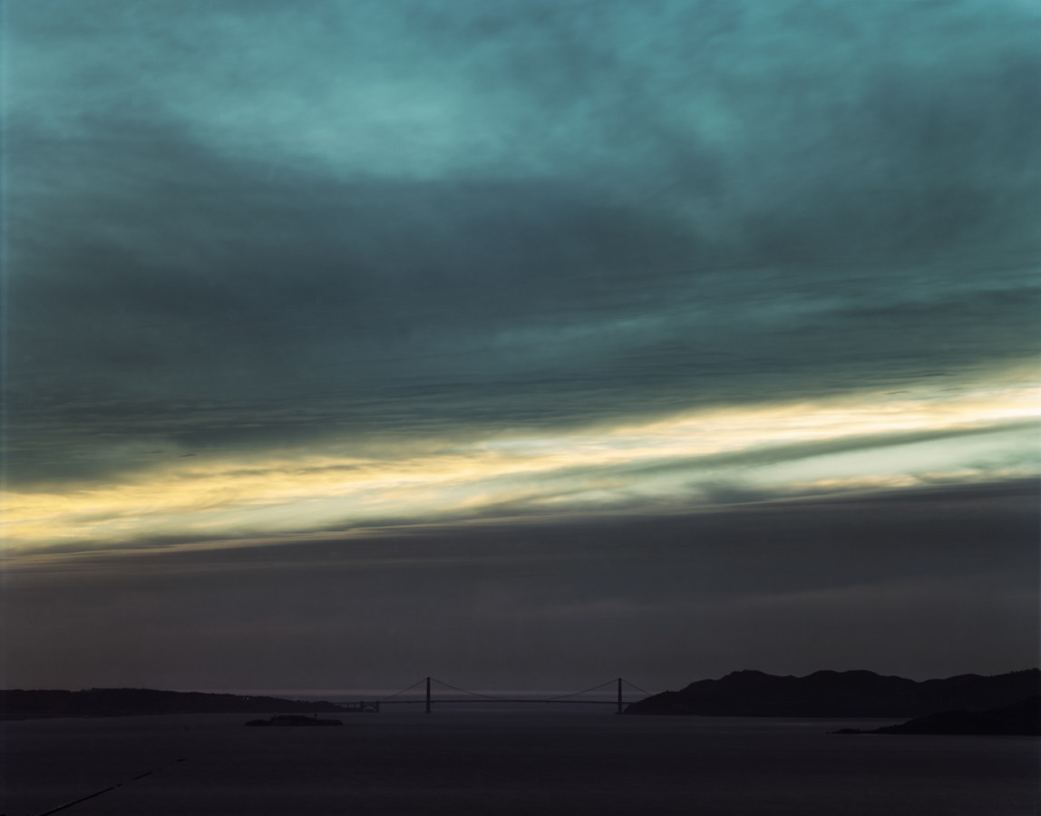 Color photograph of the distant Golden Gate Bridge under a linear break in the clouds